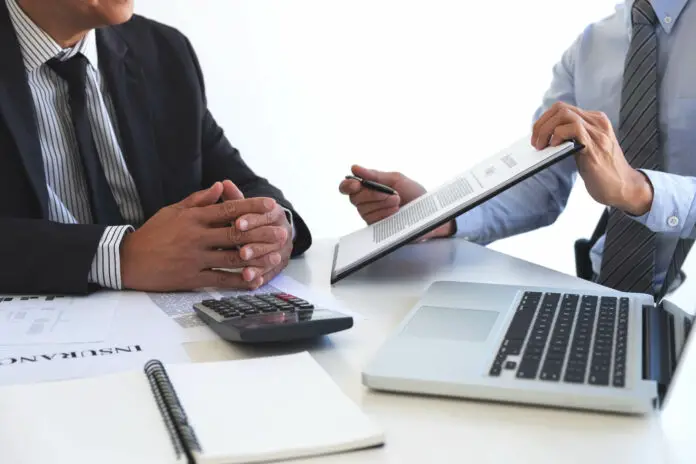 A man discussing some numbers with another on a paper and laptop